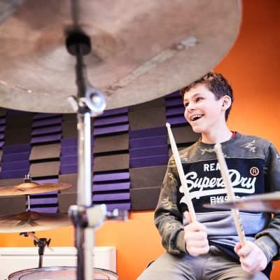 A boy wearing a Superdry jumper is playing the drums. He has a big smile on his face